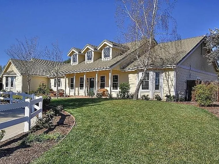 Pale yellow farmhouse style home with three dormers, a long covered front porch and a white rail fence beside a green lawn at 1448 Remington Dr, Santa Ynez, CA 93460